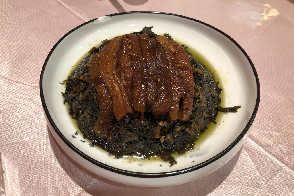 Steamed pork belly slices over dark preserved mustard greens in a white bowl