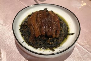 Steamed pork belly slices over dark preserved mustard greens in a white bowl