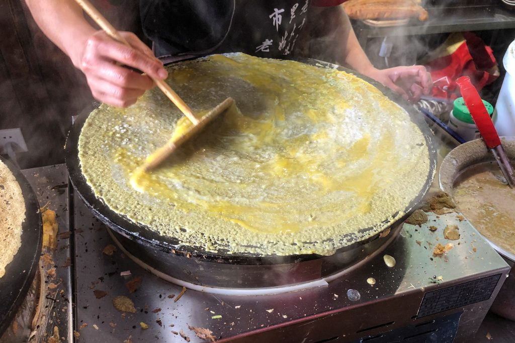 Vendor spreading egg over a thin jianbing crepe on a round griddle with fried dough sticks nearby