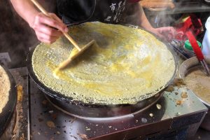 Vendor spreading egg over a thin jianbing crepe on a round griddle with fried dough sticks nearby