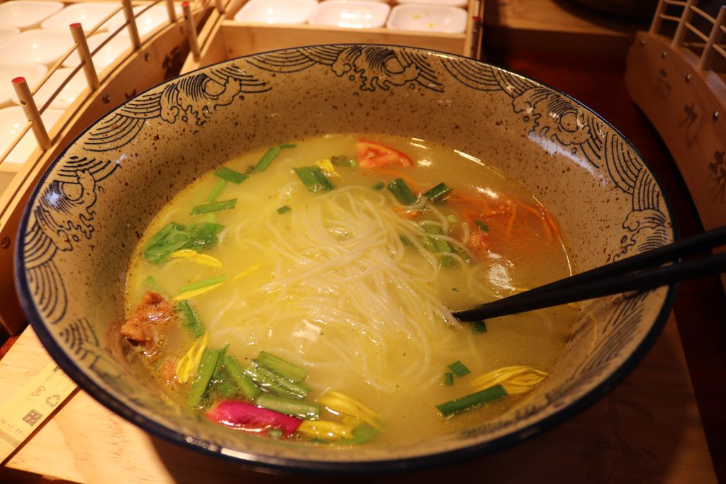 Bowl of Yunnan crossing-bridge rice noodles with broth, meat slices, and vegetable toppings