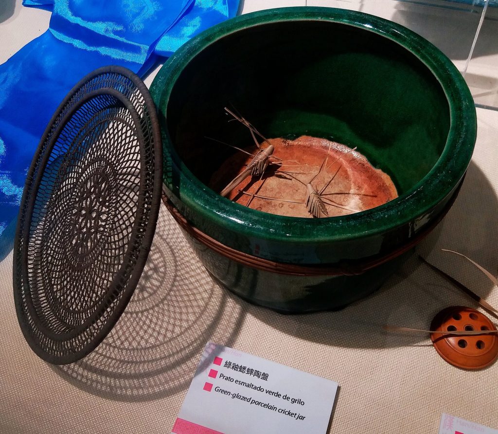 Traditional green ceramic cricket pot with a mesh lid and a cricket inside, displayed with a fighting-cricket exhibit label