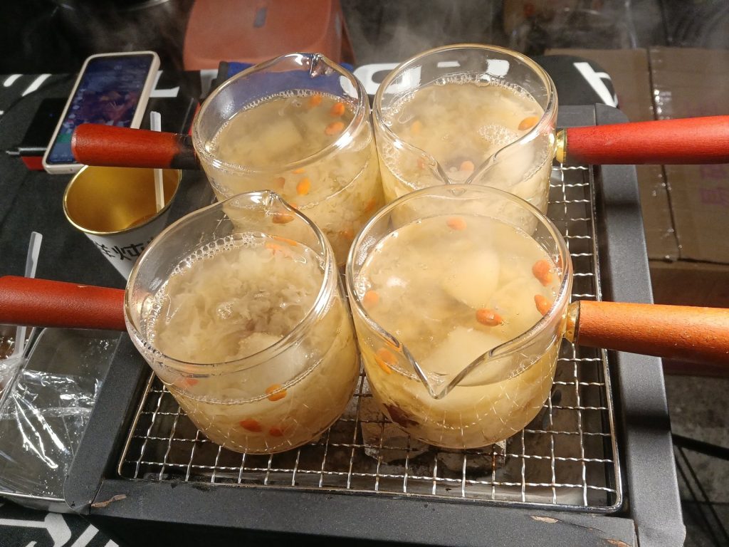 Small glass pots of rock sugar pear soup with stewed pear pieces and goji berries warming on a grill rack