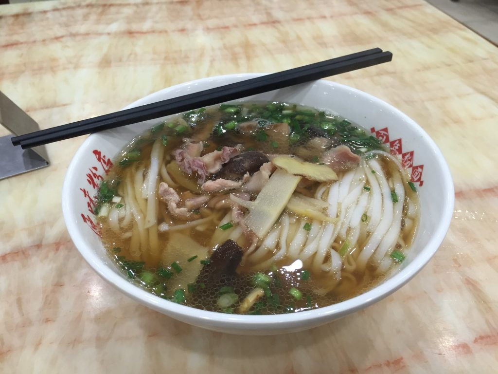 Bowl of Changsha flat rice noodles in clear broth topped with sliced meat, mushrooms, bamboo shoots and scallions