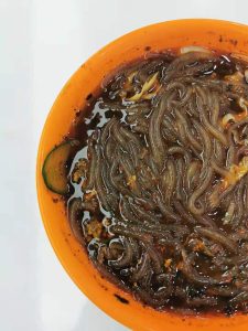 Close-up of a bowl of suanlafen with translucent sweet potato glass noodles in a dark, chili-oil and vinegar broth