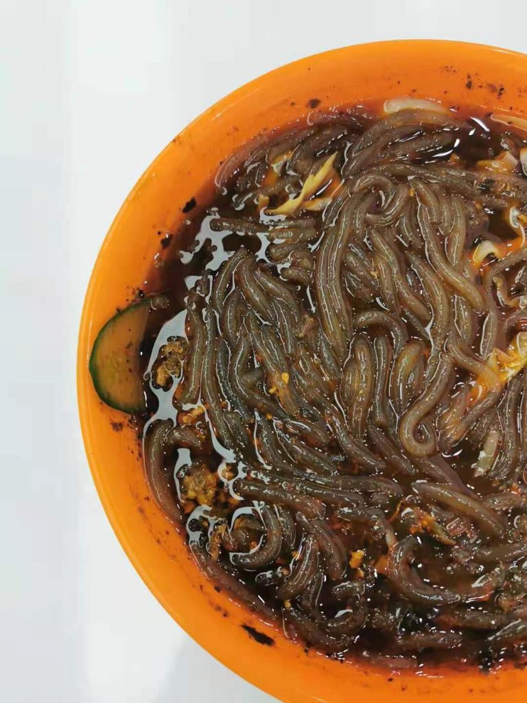 Close-up of a bowl of suanlafen with translucent sweet potato glass noodles in a dark, chili-oil and vinegar broth