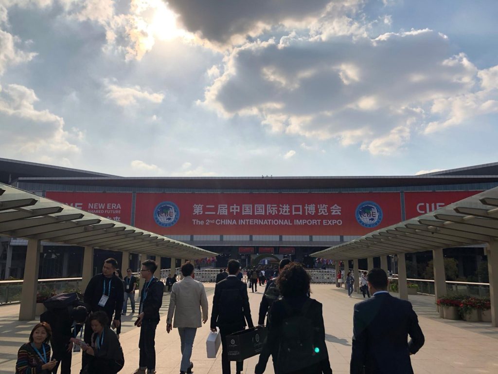 Business visitors with lanyards walking across the plaza toward the National Exhibition and Convention Center in Shanghai under a red China International Import Expo banner