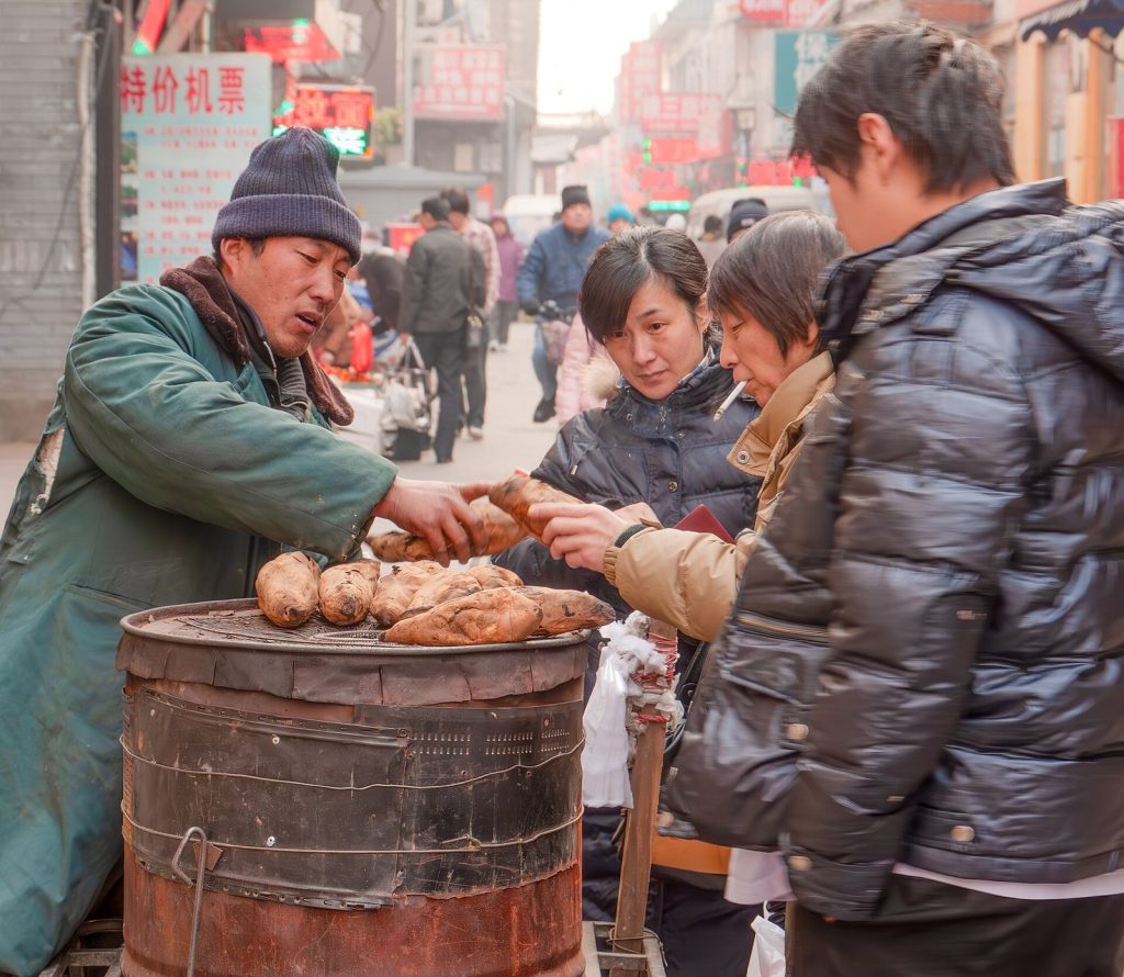 A street vendor in a green padded coat handing roasted sweet potatoes from a barrel oven to customers in winter jackets on a busy Beijing street