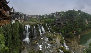 Two-tier waterfall pouring over a cliff beneath wooden stilt houses in Furong Ancient Town, Yongshun County, western Hunan