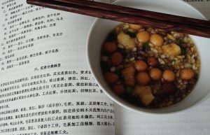 White bowl of Guangxi oil tea (youcha) topped with puffed rice, fried dough balls and chopped spring onion, with chopsticks resting on the rim