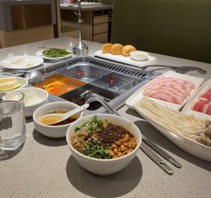 Haidilao hot pot table with a divided pot of red and clear broth, plates of sliced beef and lamb, noodles and a bowl of dipping sauce