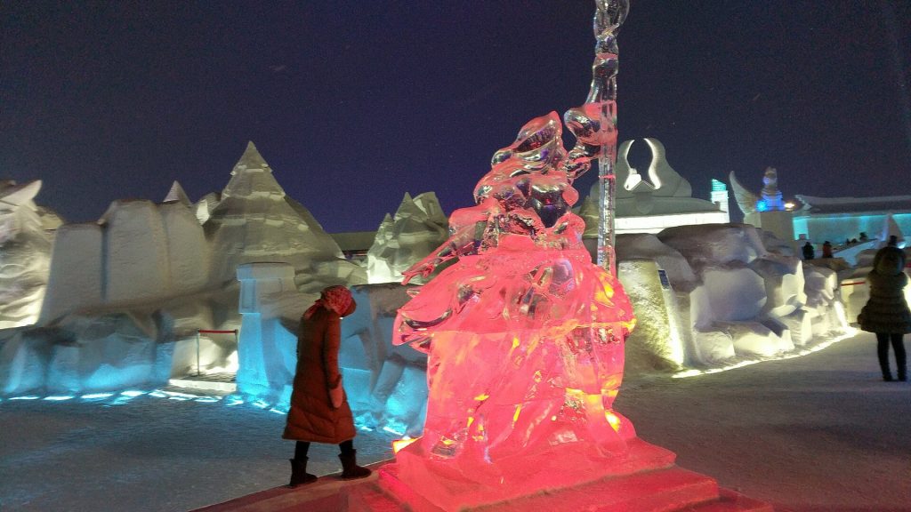 Visitor in a red coat beside a glowing pink ice sculpture, with blue-lit snow and ice buildings behind, at night in Harbin Ice and Snow World