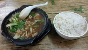 Black clay pot of huangmenji braised chicken with green peppers and greens in dark sauce, next to a bowl of white rice