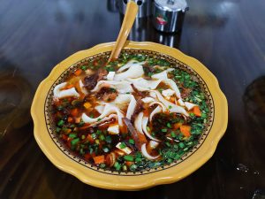Yellow bowl of Qishan saozi noodles in red sour-spicy broth with diced pork, tofu cubes, carrot, wood ear and chopped chives