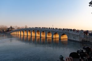 Golden winter-solstice sunset light glowing inside the arches of the Seventeen-Arch Bridge over the frozen Kunming Lake at Beijing's Summer Palace, with crowds and photographers on the shore