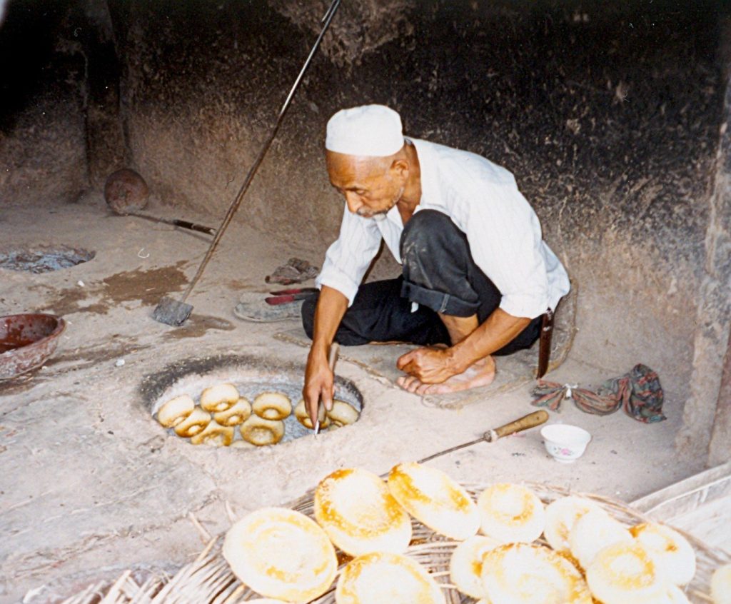 Baker crouching beside a clay tandoor oven in Kashgar with freshly baked round nang breads stacked beside him