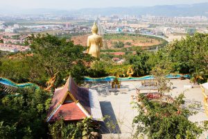 View over Jinghong city in Xishuangbanna from a hillside temple area, with a tall golden Buddha statue, Dai-style temple roofs and tropical trees in the foreground