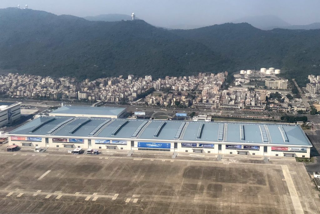 Aerial view of the long blue-roofed exhibition halls and apron of the Zhuhai International Airshow Center, with Jinwan District housing and green hills behind
